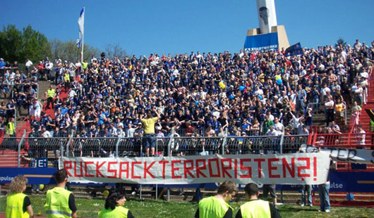 Vor dem Gästeblock hing das Transparent mit der Aufschrift „Rucksackterroristen“.<br />Bild: www.infernokoblenz.net“><figcaption>Vor dem Gästeblock hing das Transparent mit der Aufschrift „Rucksackterroristen“.<br />Bild: www.infernokoblenz.net</figcaption></figure>
<figure><img decoding=